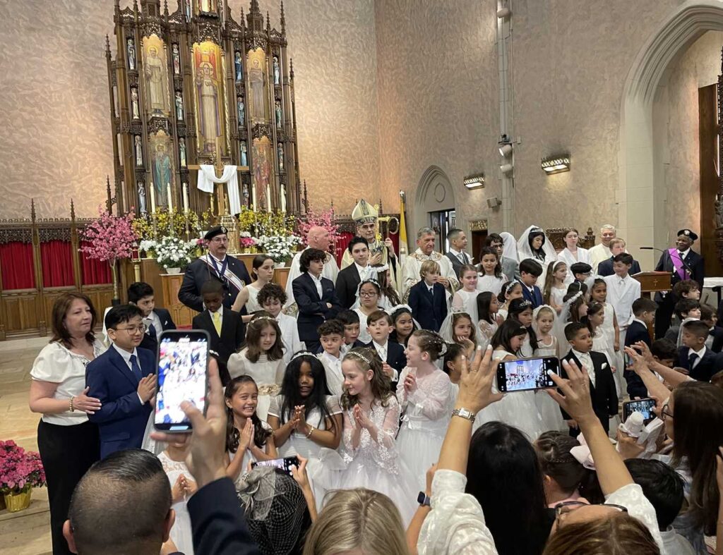 A large gathering of girls attired in white communion dresses and boys in dark suits and ties standing in front of the altar at Our Lady Queen of Martyrs Church. The altar is attired for Easter. The arms of a few paparazzi parents are shown, capturing photos on their cell phones.