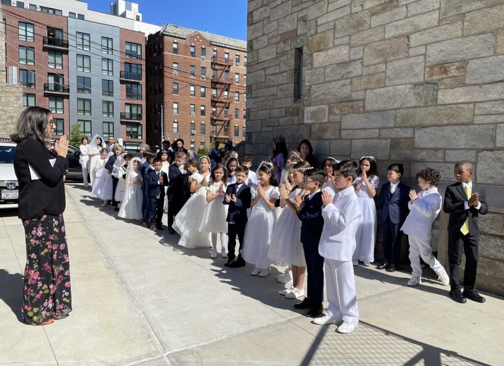 A group of girls attired in white communion dresses and boys in communion suits stand with their hands clasped together in prayer pose. Facing them is their religious education teacher also with her hands folded in prayer. They are lined up outside in bright sunlight and preparing to enter the church to receive First Holy Communion.